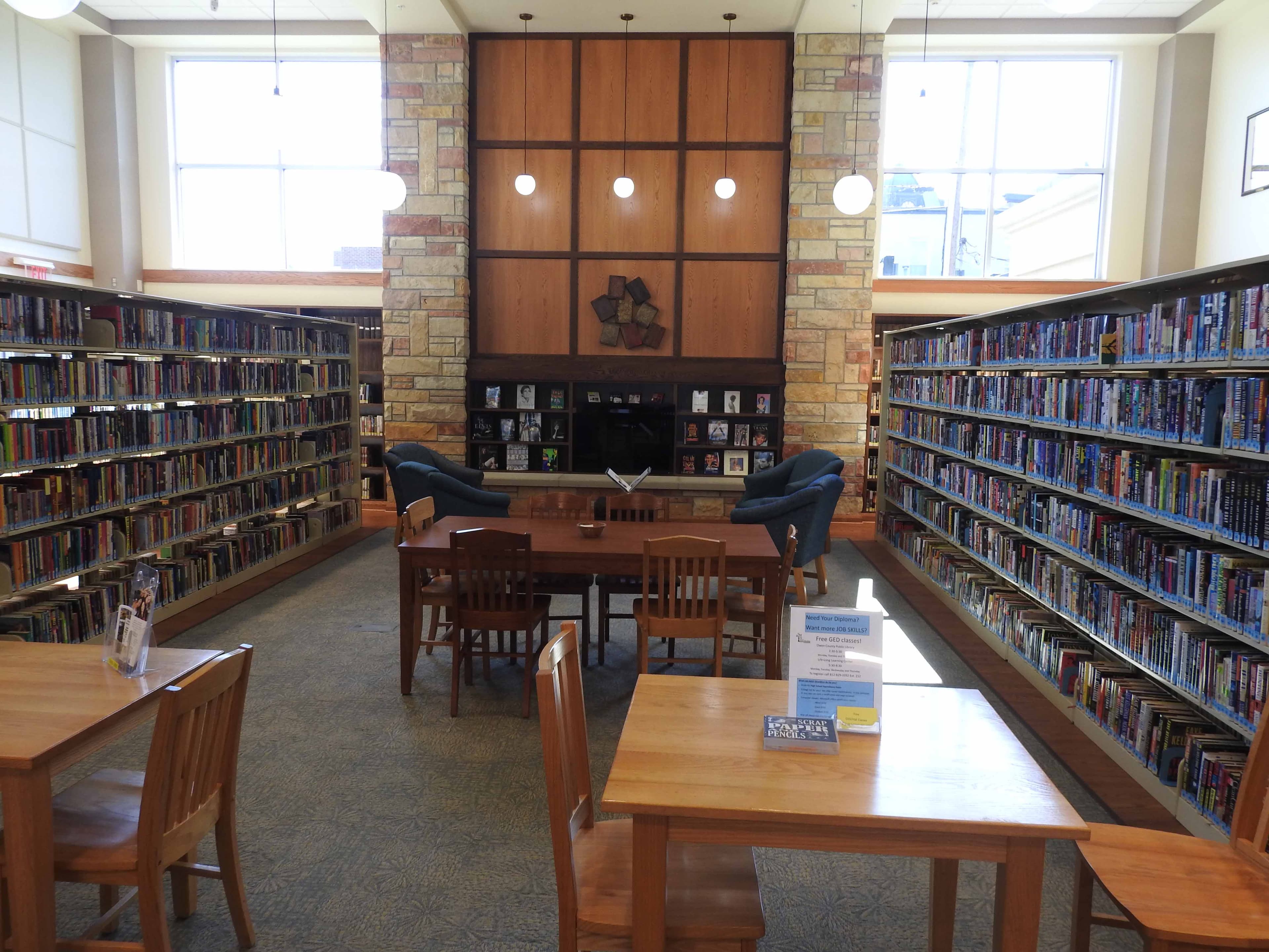 A reading area at the library with tables and chairs flanked by tall shelves of books, with a stone fireplace at the center.