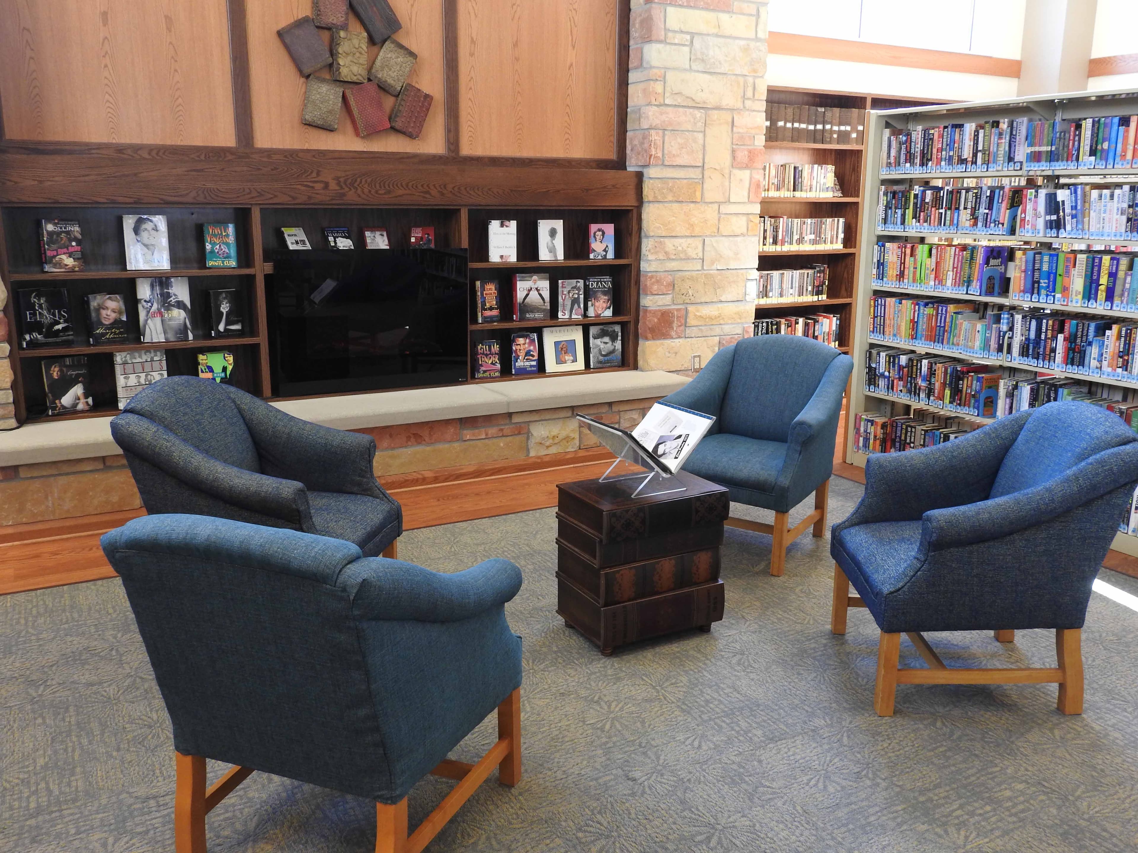 A cozy lounge area with blue armchairs arranged around a small table, opposite a stone fireplace, with display shelves of audiobooks and movies on the wall and book stacks visible to the right.